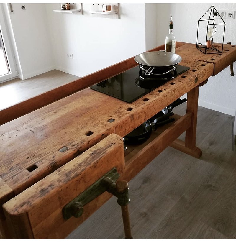 Holztisch mit Kochfeld, Küchenutensilien und einer Glaslampe auf der Oberfläche in einem modernen Raum.