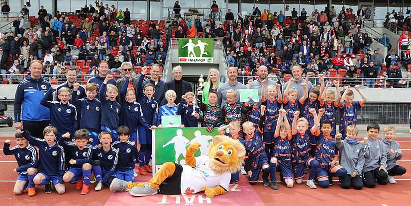 Gruppenfoto von jubelnden Kindern und Trainern nach einem Fußballturnier, mit einem Maskottchen und Pokalen im Hintergrund.