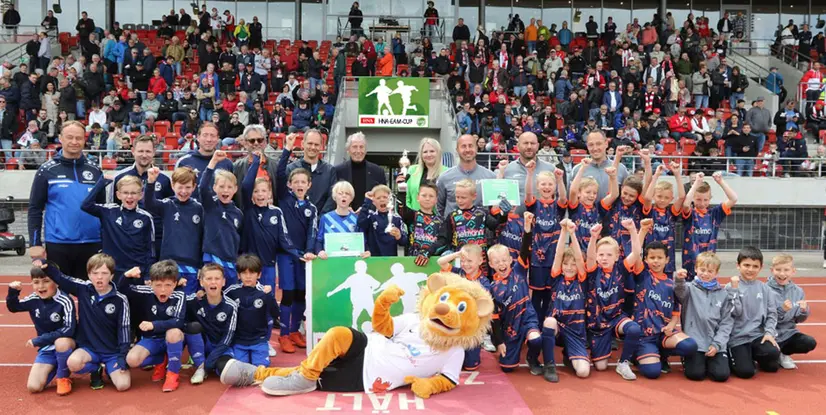 Gruppenfoto von jubelnden Kindern und Trainern nach einem Fußballturnier, mit einem Maskottchen und Pokalen im Hintergrund.