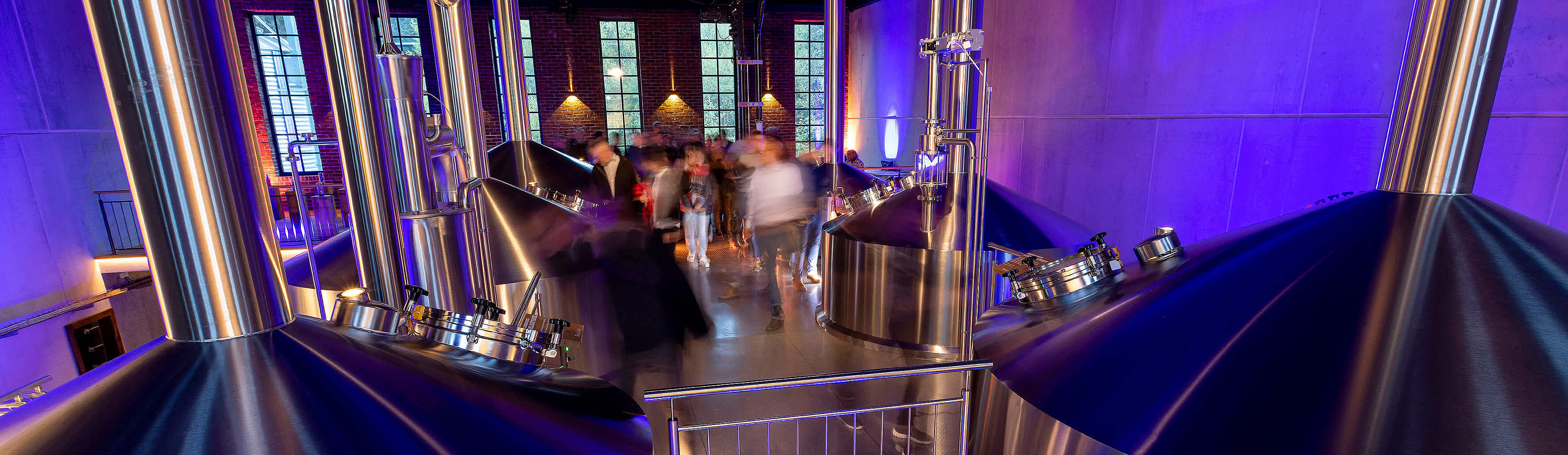 Große, glänzende Brauerei-Behälter in einem modernen Raum mit blauen und lila Lichtern, Menschen bewegen sich im Hintergrund.