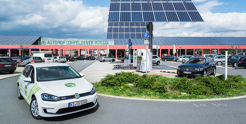 Ein Volkswagen E-Auto parkt an einer Ladestation in einem modernen Autohof mit Solarpanelen im Hintergrund.