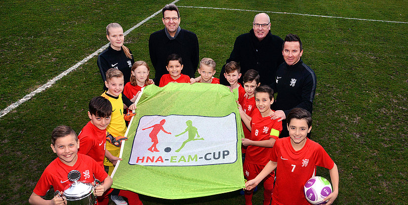 Gruppenfoto von Kindern und Trainern mit einem Banner zum HNA-EAM-Cup auf einem Fußballplatz.