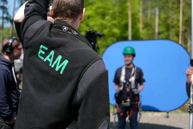 Ein Mann mit einem schwarzen Fleecejacke mit grüner Aufschrift "EAM"" steht vor einer blauen Kulisse