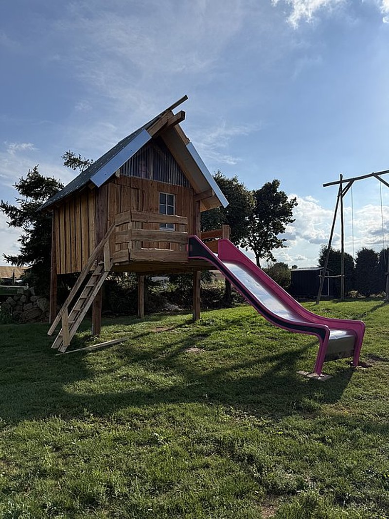 Bauernhaus mit Rutsche auf einem Spielplatz, umgeben von Wiese und Bäumen, bei sonnigem Wetter.