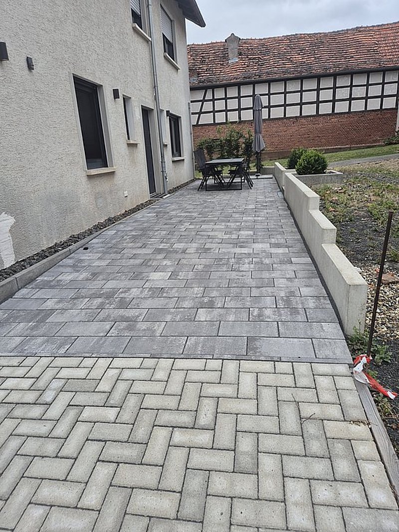 Terrasse mit grauen Pflastersteinen, Tisch und Stühlen, neben einer Wand und einer Rasenfläche. Übercast Himmel.