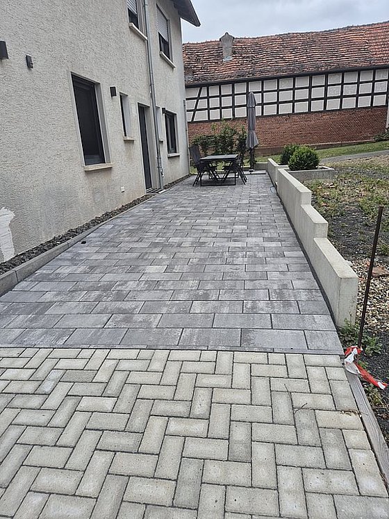 Terrasse mit grauen Pflastersteinen, Tisch und Stühlen, neben einer Wand und einer Rasenfläche. Übercast Himmel.