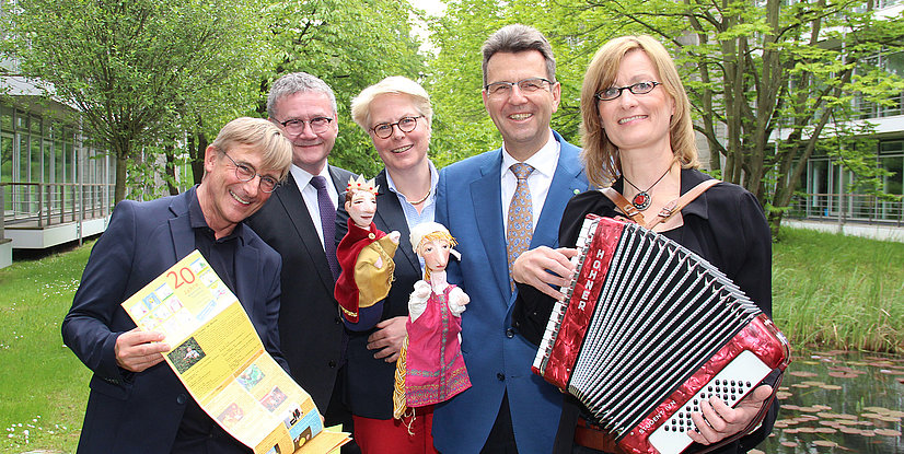 Gruppenszene mit fünf Personen, die Puppen und ein Akkordeon halten, umgeben von einem grünen Park.