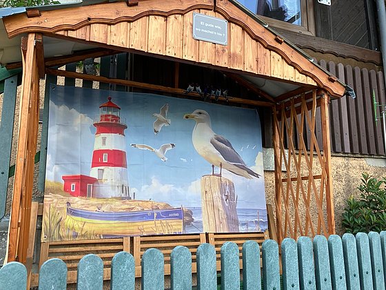 Ein hölzerner Unterstand mit einem großen Wandbild eines Leuchtturms, einer Möwe und Boote vor blauem Himmel.