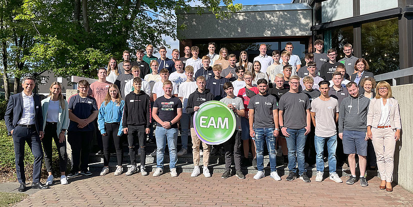 Gruppenbild von den neuen Auszubildenden, die vor einem Gebäude stehen, einige halten ein EAM-Schild. Freundliche Atmosphäre, sonniger Tag.