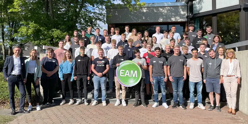 Gruppenbild von den neuen Auszubildenden, die vor einem Gebäude stehen, einige halten ein EAM-Schild. Freundliche Atmosphäre, sonniger Tag.