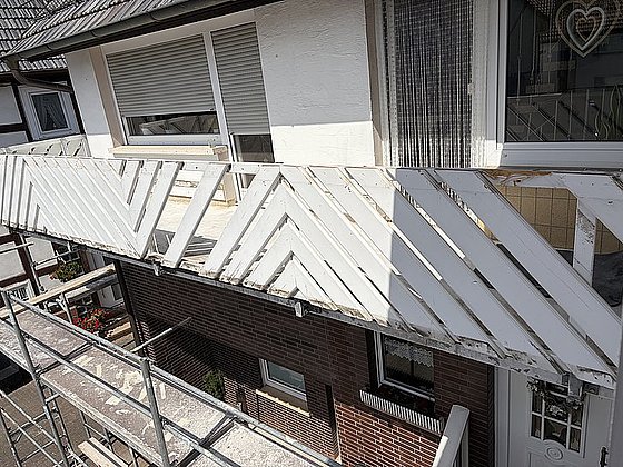 Balkon mit weißen Holzlatten und Gerüst, Blick auf das Obergeschoss eines Hauses mit Fenster und Blumen.