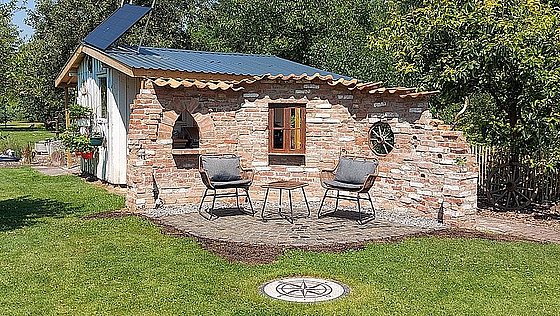 Gemütlicher Gartenbereich mit Steinhütte, zwei Stühlen und einem Tisch auf Pflastersteinen, umgeben von Grün.