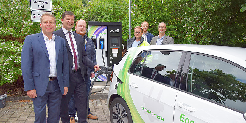 Personen stehen an einer öffentlichen Ladestation für Elektroautos, während ein Auto aufgeladen wird.