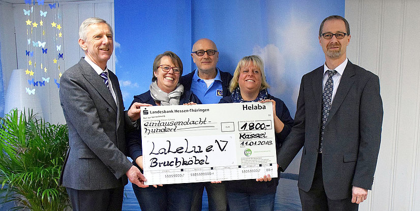 Gruppe von fünf Personen hält einen Scheck über 1.800 Euro für den Verein LaLeLu e.V. vor einer blauen Wand.