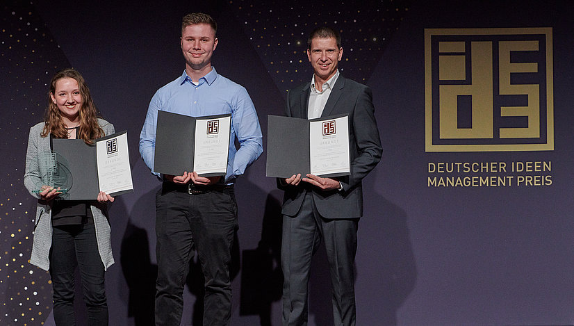 Drei Personen stehen auf einer Bühne, präsentieren Urkunden und einen Preis beim Deutschen Ideen Management Preis.