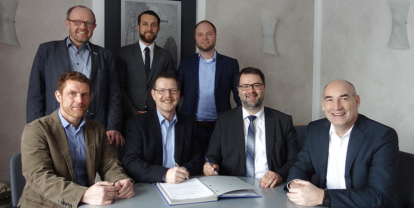 Gruppenselbstporträt mit mehreren Männern, die um einen Tisch versammelt sind, während sie Dokumente unterschreiben.