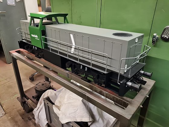 Modell einer Lokomotive in grüner und grauer Farbgebung, abgestellt auf einer Werkbank vor grüner Wand.