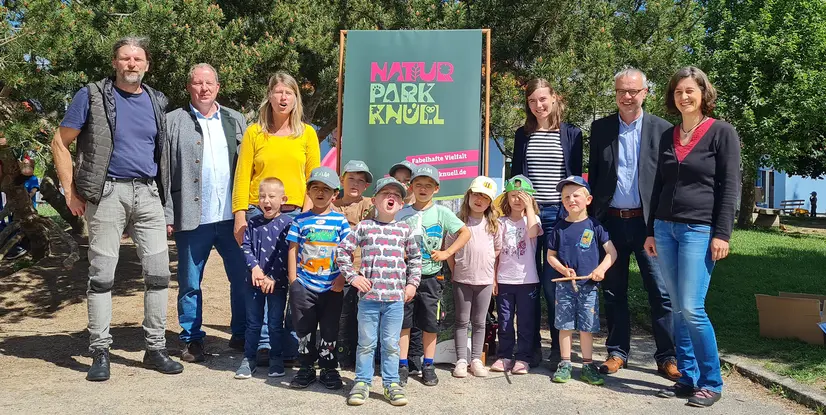 Gruppe von Erwachsenen und Kindern vor einem Naturpark-Schild, lächelnd im Freien, umgeben von Bäumen und Wiese.