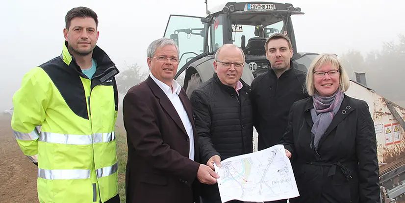 Fünf Personen stehen in Arbeitskleidung vor einem Traktor und halten einen Plan in der Hand, Umgebungsnebel sichtbar.