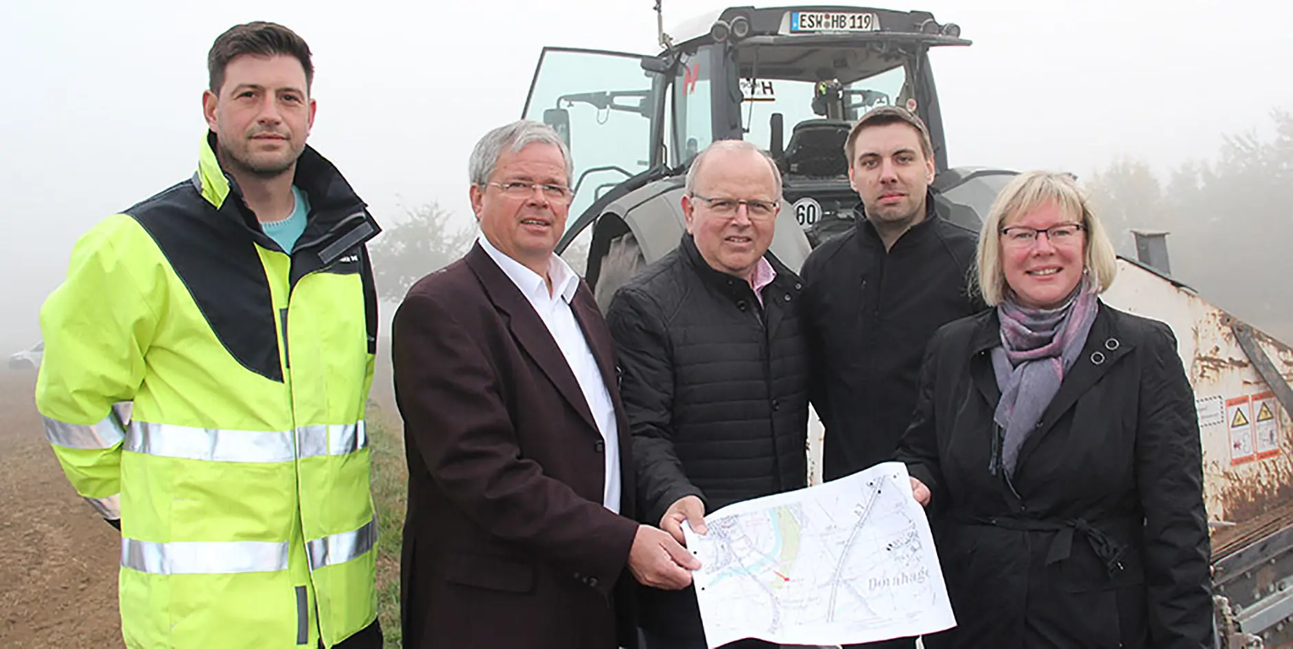 Fünf Personen stehen in Arbeitskleidung vor einem Traktor und halten einen Plan in der Hand, Umgebungsnebel sichtbar.