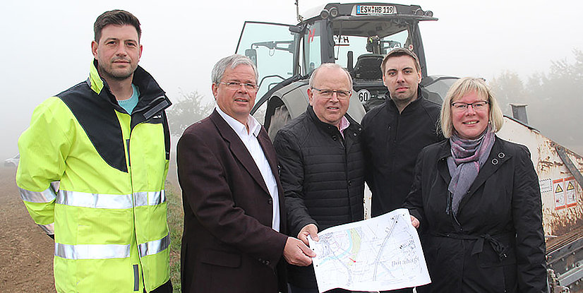 Fünf Personen stehen in Arbeitskleidung vor einem Traktor und halten einen Plan in der Hand, Umgebungsnebel sichtbar.