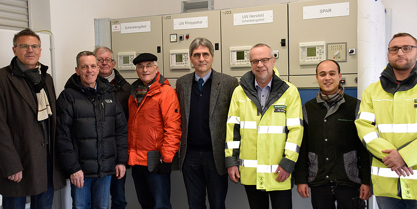 Gruppe von sieben Personen in Arbeitskleidung vor elektrischen Schaltschränken in einem technischen Umfeld.