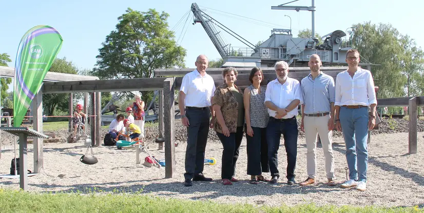 Gruppenszene auf einem Spielplatz mit sechs Personen, einem grünen Banner und spielenden Kindern im Hintergrund.