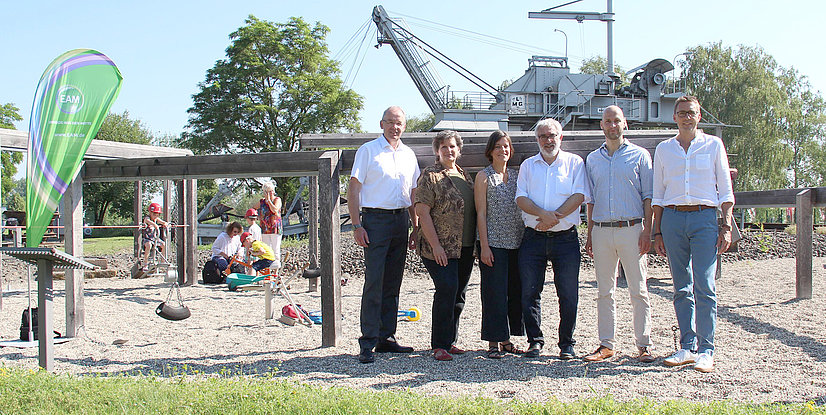 Gruppenszene auf einem Spielplatz mit sechs Personen, einem grünen Banner und spielenden Kindern im Hintergrund.