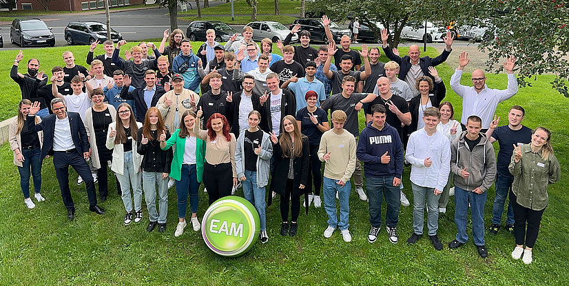 Großes Gruppenfoto, die im Freien stehen und zum Fotografen winken oder Daumen hoch zeigen. EAM-Logo im Vordergrund.