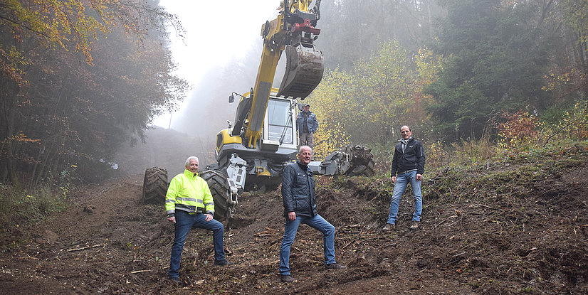 Vier Männer stehen vor einem Bagger in einem nebligen Waldgebiet mit herbstlichen Bäumen im Hintergrund.