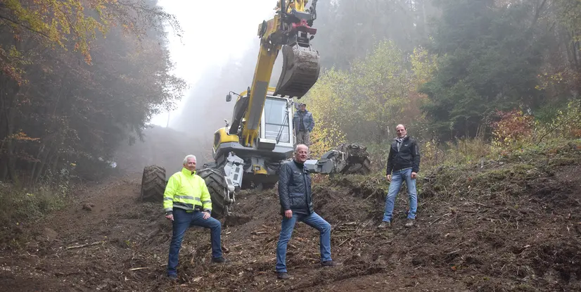 Vier Männer stehen vor einem Bagger in einem nebligen Waldgebiet mit herbstlichen Bäumen im Hintergrund.