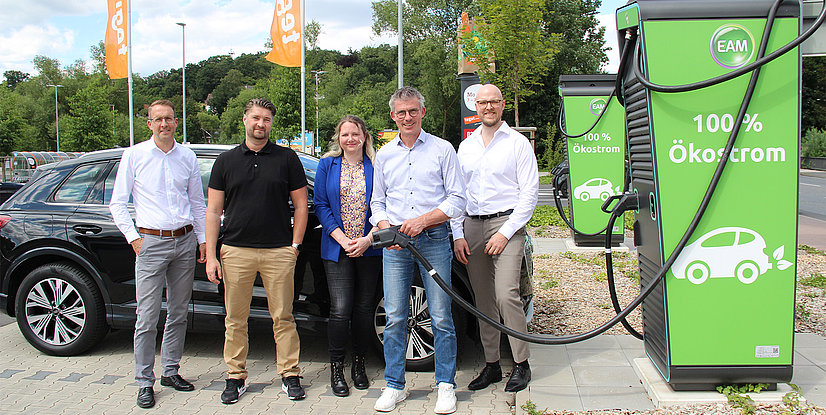Fünf Personen stehen vor einer Elektroauto-Ladestation mit 100 % Ökostrom auf grünem Hintergrund.