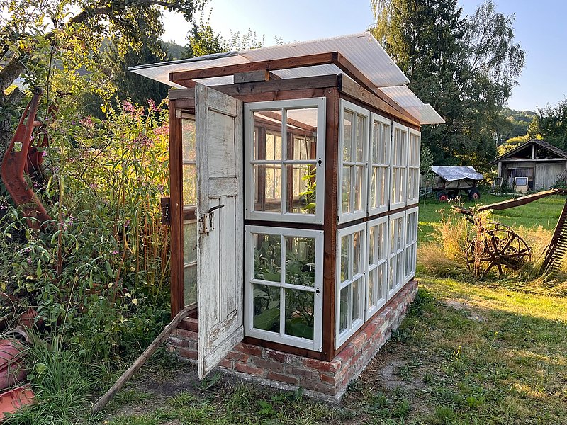 Ein Gartenhaus mit großen Fenstern, offenem Eingang und umgeben von bunten Blumen und Pflanzen im Vordergrund.