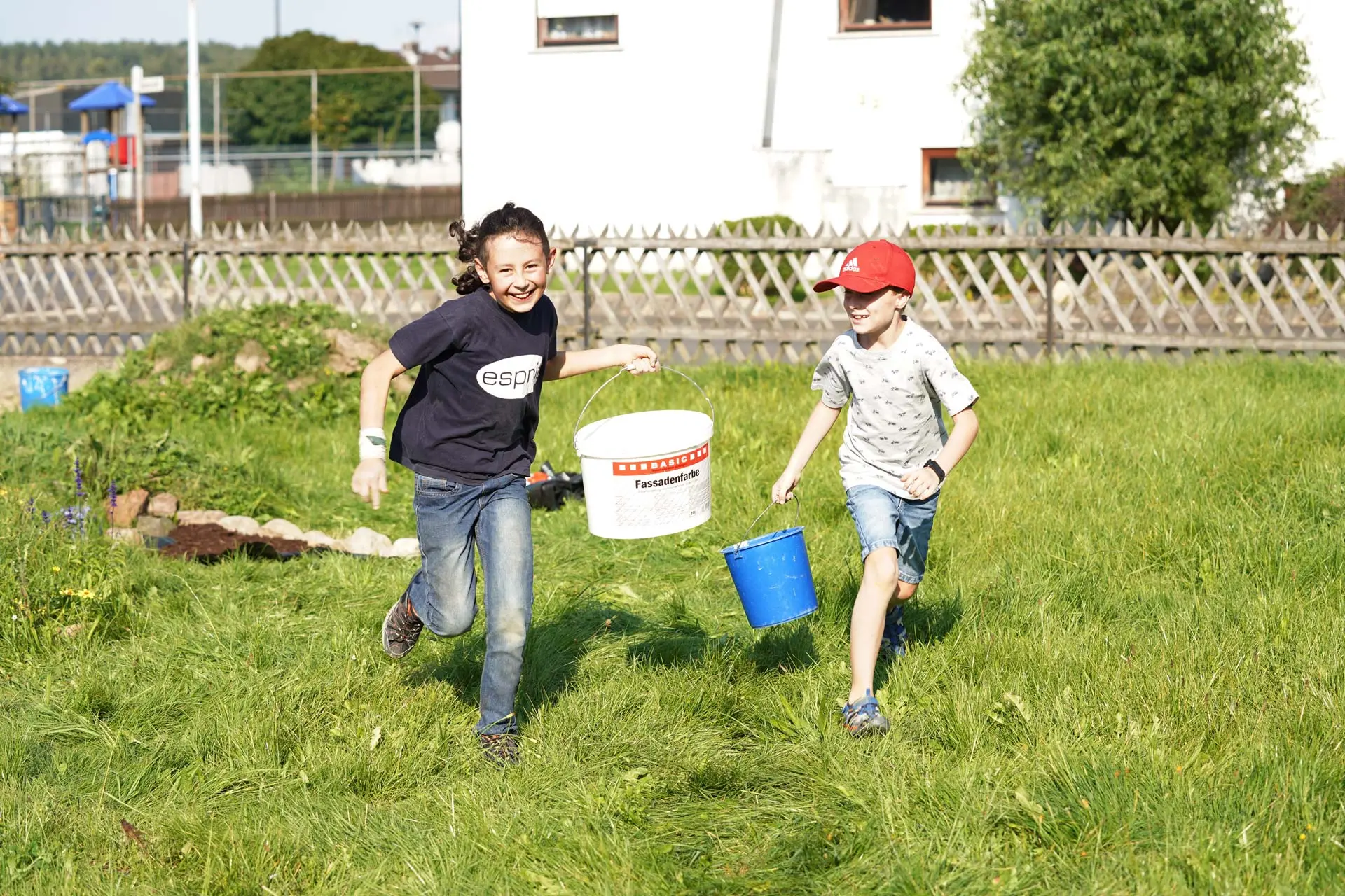 Zwei Kinder laufen fröhlich über eine Wiese und tragen Eimer in den Händen. Die Sonne scheint.