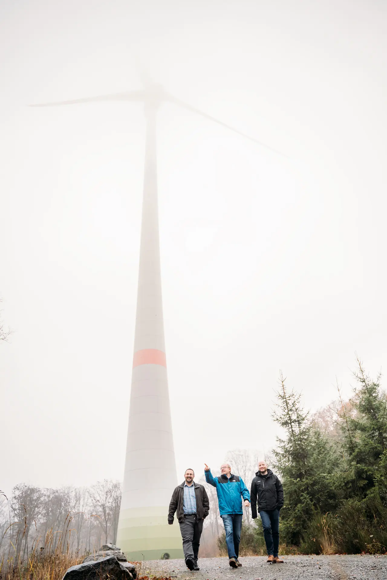 Drei Männer gehen auf einem Weg neben einem Windrad, umgeben von Nebel und Bäumen.