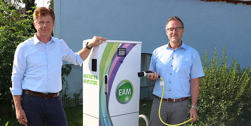 Zwei Männer stehen neben einer E-Ladesäule, einer hält ein Ladekabel, umgeben von Pflanzen.