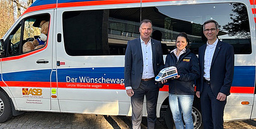 Die EAM-Geschäftsführer Olaf Kieser und Hans-Hinrich Schriever übergeben die Spende an die Projektkoordinatorin des Wünschewagens Nordhessen Larissa Groffmann vor dem Wünschewagen, an dessen Steuer ein riesiger Teddy sitzt.