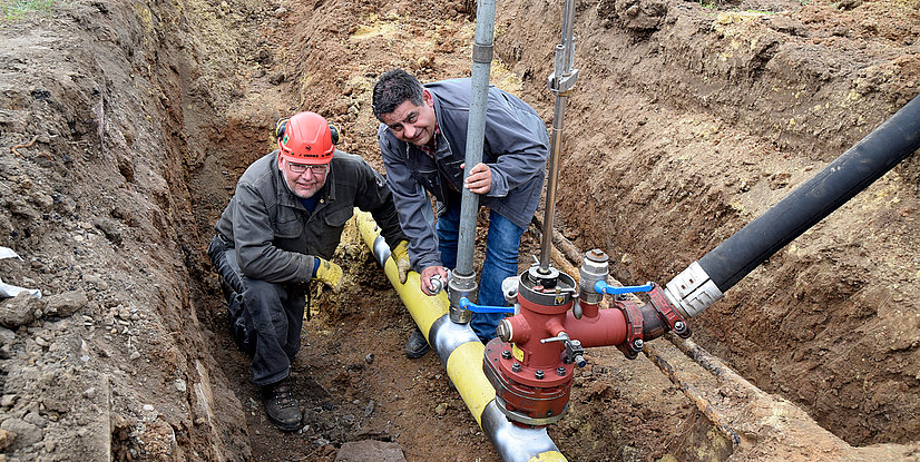 Zwei Arbeiter in einem Graben, die an einem Rohrleitungssystem mit Ventilen und Schläuchen arbeiten.