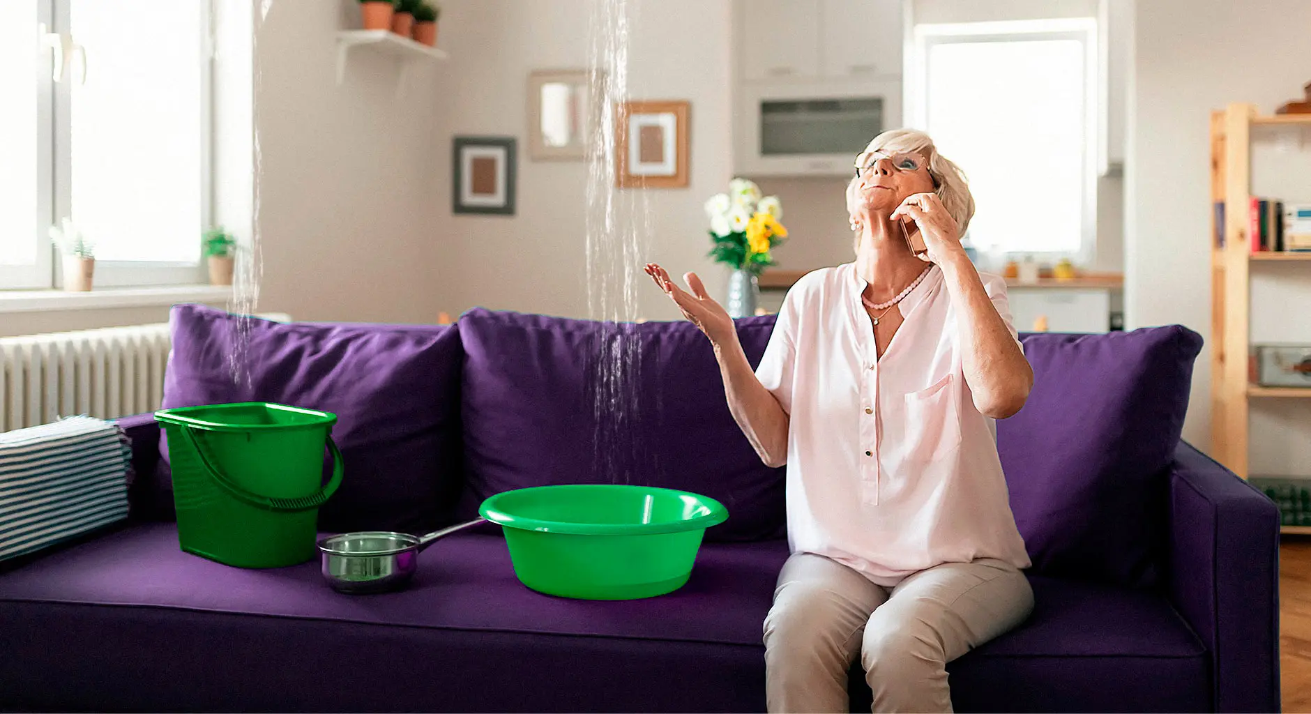 Eine ältere Frau sitzt auf einer lila Couch, schaut nach oben und spricht besorgt telefonierend, während Wasser von der Decke tropft.