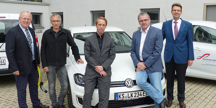 Fünf Männer posieren neben einem weißen VW E-Golf mit EAM-Branding und einem EAM-Fahrzeug im Hintergrund.
