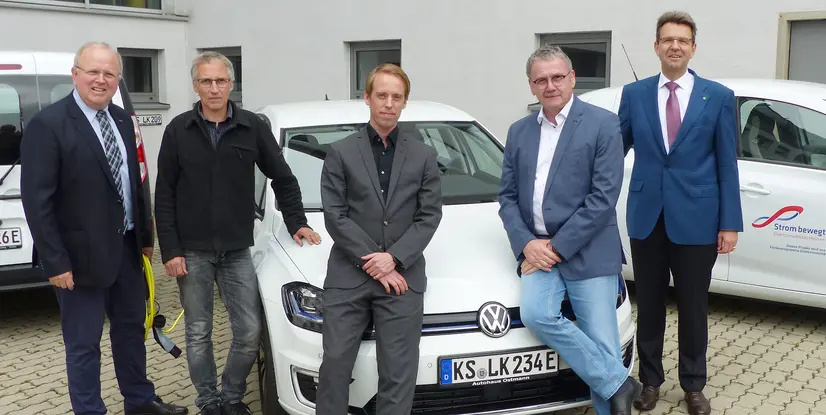 Fünf Männer posieren neben einem weißen VW E-Golf mit EAM-Branding und einem EAM-Fahrzeug im Hintergrund.