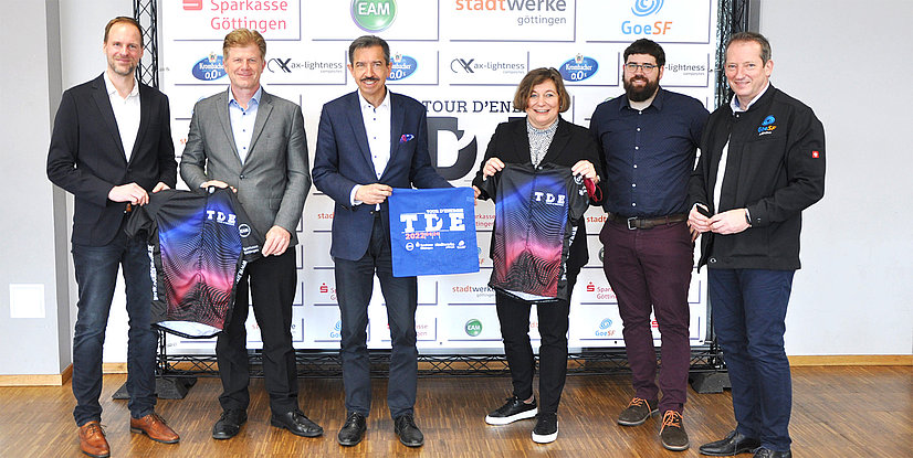 Gruppenbild von sechs Personen bei einer Veranstaltung, die T-Shirts und ein Banner für die Tour d'Energie präsentieren.