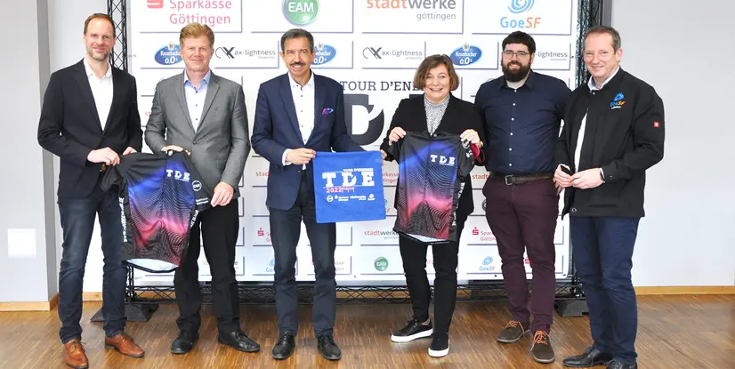 Gruppenbild von sechs Personen bei einer Veranstaltung, die T-Shirts und ein Banner für die Tour d'Energie präsentieren.