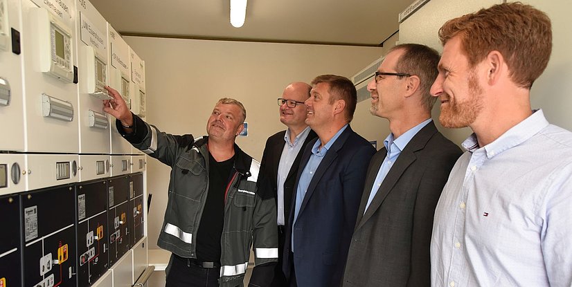 Fünf Männer stehen vor elektrischen Messtechnik-Geräten und diskutieren, während einer auf ein Gerät zeigt.