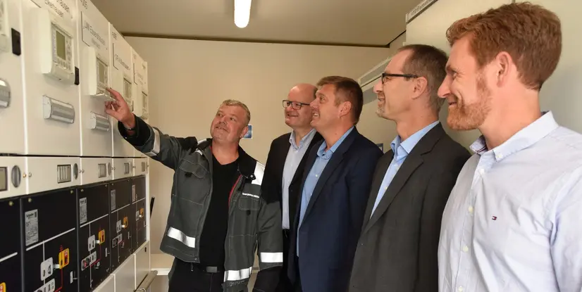 Fünf Männer stehen vor elektrischen Messtechnik-Geräten und diskutieren, während einer auf ein Gerät zeigt.