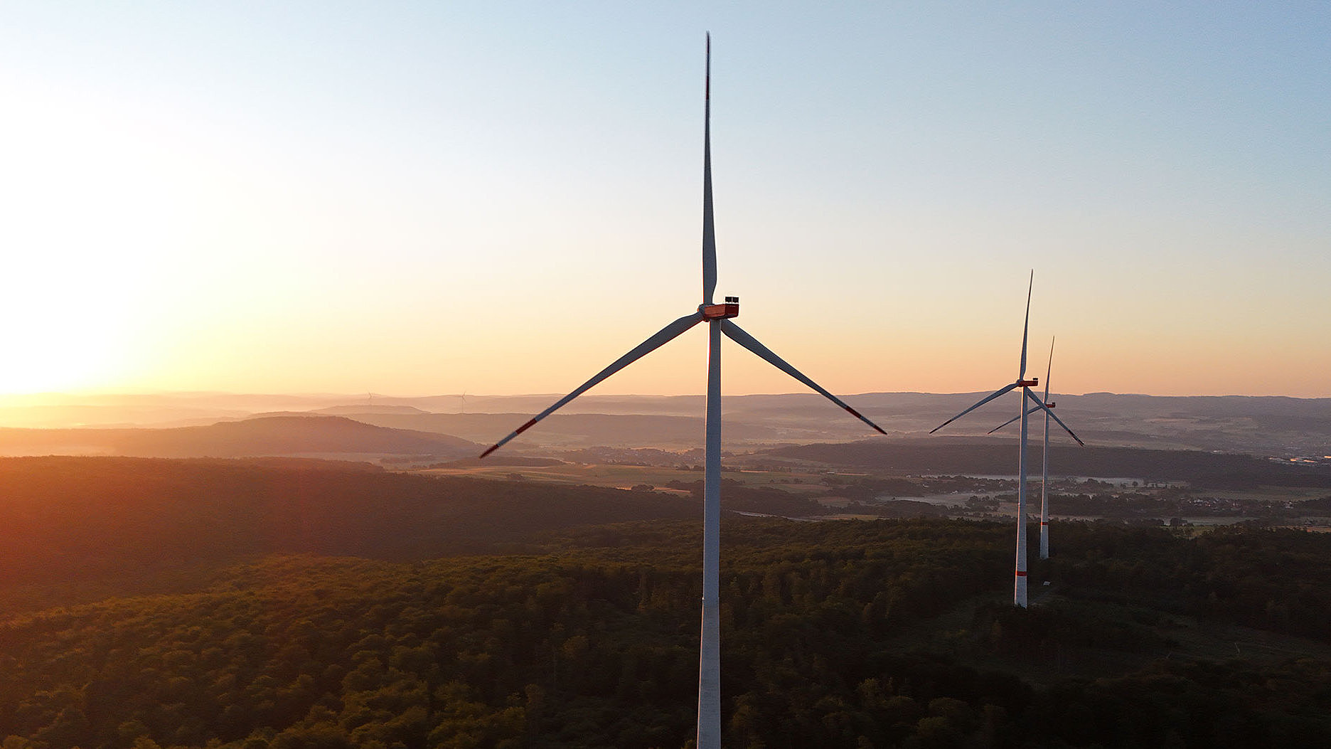 Zwei Windturbinen stehen im Vordergrund, vor einer goldenen Abenddämmerung über einer bewaldeten Landschaft.