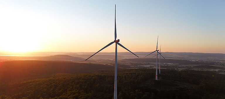 Zwei Windturbinen stehen im Vordergrund, vor einer goldenen Abenddämmerung über einer bewaldeten Landschaft.