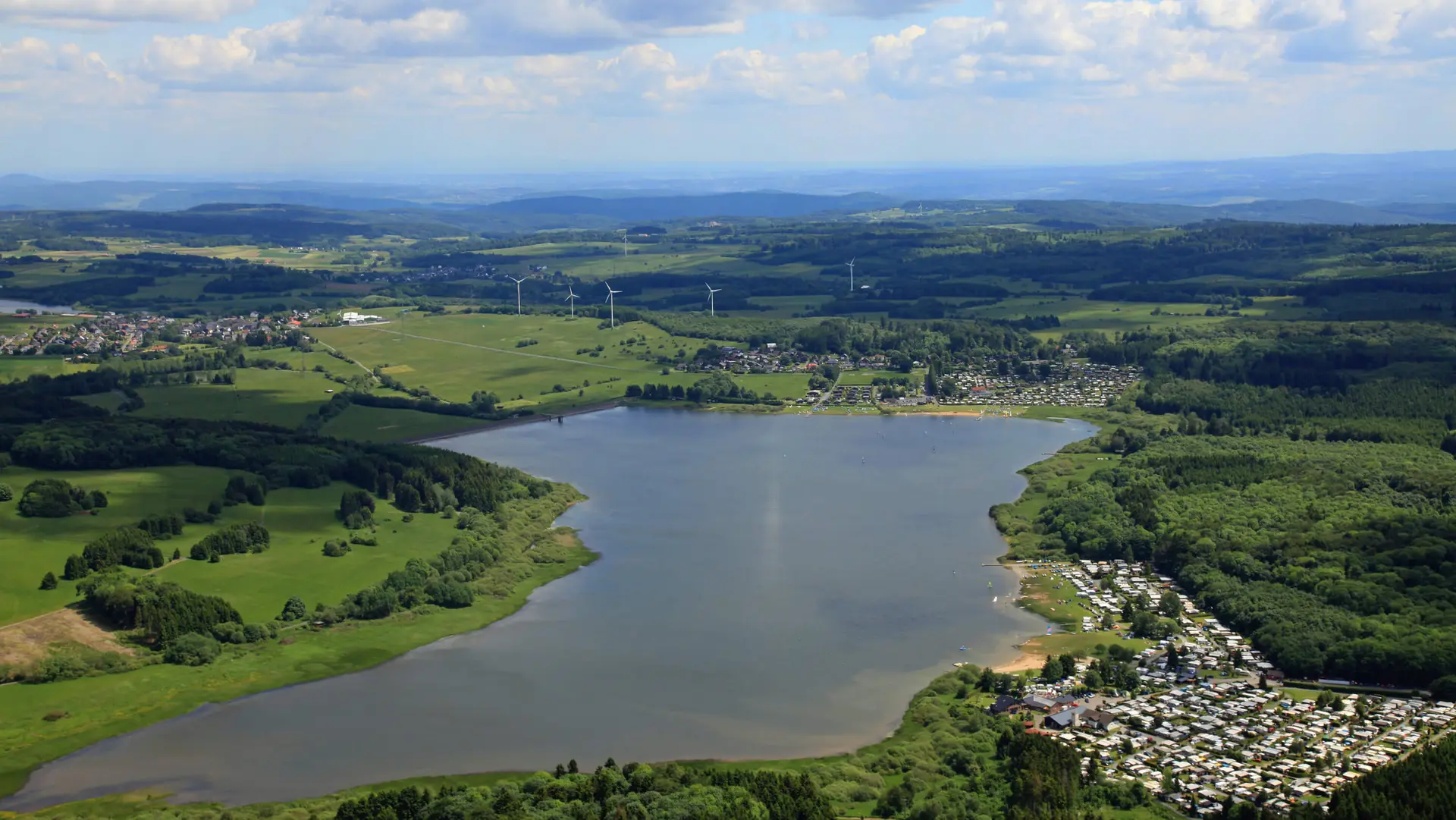 Blick von oben auf die Krombachtalsperre, umgeben von grünen Wäldern, Wiesen, einem Campingplatz und Windrädern am Horizont unter blauem Himmel.