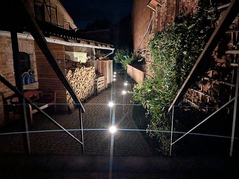 Ein beleuchteter Weg mit Steinpflaster, umgeben von Zäunen, Holzstapeln und Pflanzen, bei Nacht aufgenommen.
