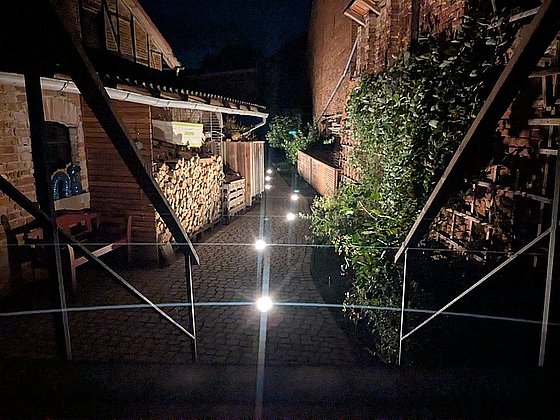 Ein beleuchteter Weg mit Steinpflaster, umgeben von Zäunen, Holzstapeln und Pflanzen, bei Nacht aufgenommen.